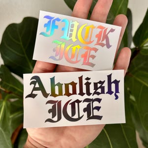 May include: Two stickers with text. The top sticker reads "FUCK ICE" in rainbow holographic lettering. The bottom sticker reads "Abolish ICE" in black and holographic lettering. Both stickers are rectangular and have a white background.