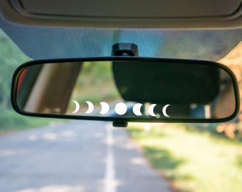 Puede incluir: Un espejo retrovisor de coche con una calcomanía blanca de las fases del ciclo lunar, incluyendo formas de cuarto creciente, media luna y luna llena, aplicada horizontalmente en el centro de la superficie de cristal.