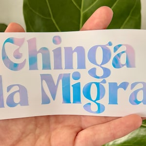 May include: A white sticker with the text "Chinga la Migra" in a blue and pink tie-dye font. The sticker is held in a hand in front of a green leafy background.