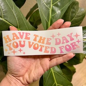 May include: A white sticker with the text "Have the day you voted for" in pink and orange letters with a gradient effect. The letters are in a retro font style. There are small pink stars around the text.