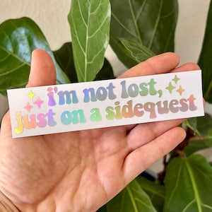 May include: A rectangular sticker with the text "I'm not lost, just on a sidequest" in holographic, pastel-colored lettering. The sticker also features star and sparkle graphics. The sticker is held in a hand in front of a green leafy background.