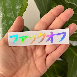 May include: A rectangular sticker with Japanese characters in a gradient of colours, including blue, green, yellow, and purple. The sticker is on a white background.