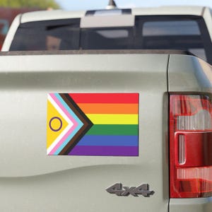 Pode incluir: Um carro com um adesivo de bandeira do orgulho na parte de trás. A bandeira é um arco-íris com um triângulo amarelo e um círculo roxo no canto superior esquerdo.