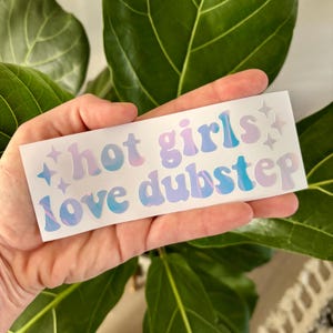 Peut inclure: Un autocollant rectangulaire avec le texte "hot girls love dubstep" dans une police rétro. Le texte est dans des tons de rose, de bleu et de violet avec des étoiles. L'autocollant est tenu devant un fond de feuilles vertes.