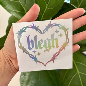 May include: A white sticker with a heart-shaped design. The heart is formed by a barbed wire outline with a rainbow holographic effect. The word "blegh" is written in a gothic font inside the heart, with four star shapes.
