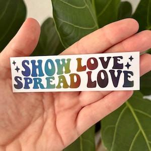 Può includere: Un adesivo rettangolare con le parole "SHOW LOVE SPREAD LOVE" in lettere a bolle color arcobaleno. L'adesivo ha uno sfondo bianco ed è decorato con piccole forme di stella.