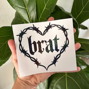 Peut inclure: Un autocollant blanc avec le mot "brat" dans une police sombre et scintillante, encadré par un motif de fil de fer barbelé en forme de cœur. L'autocollant est tenu dans une main, avec une plante verte feuillue en arrière-plan.