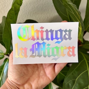 May include: A white sticker with the text "Chinga la Migra" in a holographic, rainbow-colored font. The sticker is held in a hand, with a green plant in the background.