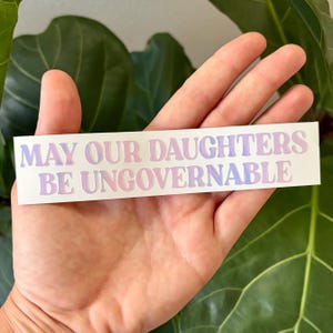以下が含まれることがあります： ピンクと青のグラデーションで「MAY OUR DAUGHTERS BE UNGOVERNABLE」と書かれた白いステッカー。ステッカーは手で持たれており、背景には緑の葉があります。