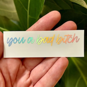 May include: A rectangular sticker with the text "you a bad bitch" in a rainbow holographic font. The sticker is held in a hand, with a green leafy background.