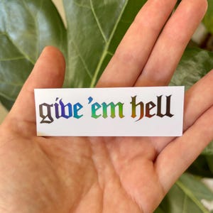 May include: A rectangular white sticker with the phrase "give 'em hell" in a gothic font. The text is in a rainbow gradient of colors, including blue, green, and red. The sticker is held in a hand.