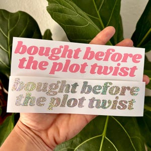 May include: Two white stickers with the text "bought before the plot twist" in pink and iridescent glitter lettering.