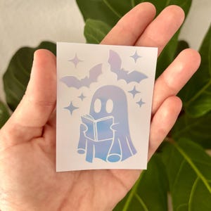 May include: A holographic sticker featuring a ghost reading a book, with bats and stars. The ghost is depicted in shades of blue and purple, with a book in its hands. The bats and stars are also in shades of purple.
