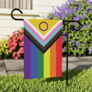 May include: A colorful garden flag featuring the Intersex-Inclusive Pride flag. The flag has a yellow top with a purple circle, and a rainbow gradient with black, brown, pink, and light blue stripes. The flag is displayed on a black metal stand.