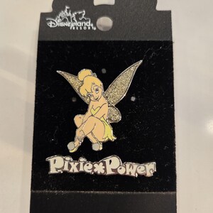 Könnte beinhalten: Ein Disney-Pin mit Tinkerbell mit glitzernden Flügeln, sitzend auf einem schwarzen Hintergrund. Der Pin trägt die Worte "Pixie Power" in Silber. Das Disneyland Resort-Logo befindet sich oben.