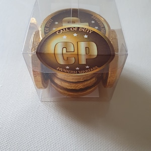 May include: A clear plastic box filled with gold-wrapped chocolate coins. Each coin has a "Call of Duty Modern Warfare" logo with "CP" lettering. The box is on a white surface. The coins are a fun treat.