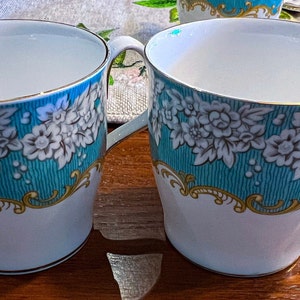 May include: Two white ceramic mugs with gold trim and a blue and white floral design. The mugs have a handle on the right side.