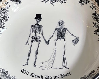May include: A white ceramic bowl with a black and white floral design. The bowl features a skeleton couple holding hands with the text "Till Death Do Us Part".