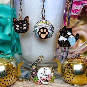 Puede incluir: Una colección de accesorios de té caprichosos. Incluye infusores de té dorados con forma de tetera y amuletos con temática de gatos. Los amuletos presentan diseños como un gato negro con múltiples ojos, una huella de pata y un gato blanco durmiendo en una luna creciente.