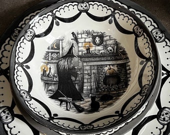 May include: A set of two black and white ceramic dinner plates with a Halloween theme. The smaller plate features a witch stirring a cauldron in a fireplace with a black cat and bats. The larger plate has a scalloped edge with a repeating pattern of bats and pumpkins.
