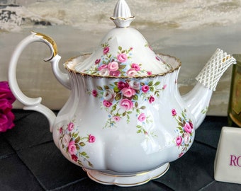 Pode incluir: Bule de porcelana branca com padrão floral de rosas cor-de-rosa e detalhes dourados. O bule tem uma alça curva e uma tampa com um botão decorativo. Um pequeno bloco retangular com "ROYAL ALBERT ENGLAND" está em primeiro plano.