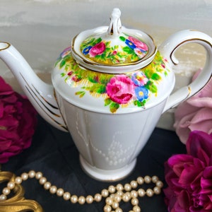 Könnte beinhalten: Eine weiße Porzellan-Teekanne mit einem Blumendesign mit rosa, blauen und gelben Blumen. Die Teekanne hat einen goldenen Rand und einen Deckel mit einem Knopf.