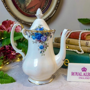 Może przedstawiać: Biały porcelanowy imbryk z niebieskim wzorem róż i złotymi obramowaniami. Imbryk ma zakrzywiony uchwyt i pokrywkę z guzikiem. Z przodu imbryka znajduje się znak Royal Albert England.