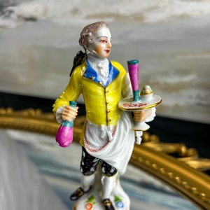 Könnte beinhalten: Porzellanfigur eines Mannes in Kleidung des 18. Jahrhunderts. Er trägt eine gelbe Jacke mit blauem Besatz, ein weißes Hemd und eine schwarze Hose. Er hält eine Flasche und ein Tablett mit einem kleinen Dessert. Die Figur steht auf einem dekorativen Sockel.