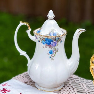 Może przedstawiać: Biały porcelanowy imbryk z pokrywką, ozdobiony niebieskimi i fioletowymi wzorami kwiatowymi róż i złotymi obramowaniami. Imbryk ma zakrzywiony uchwyt i dzióbek. Stoi na białym obrusie z czerwonym haftem kwiatowym.