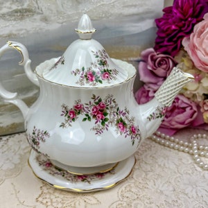 Może przedstawiać: Biały czajnik z kwiatowym wzorem i złotą obwódką. Czajnik ma pokrywkę i uchwyt. Stoi na białej podstawce.