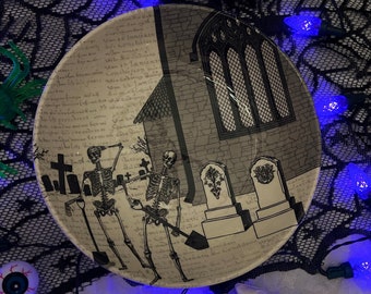 May include: A round, Halloween-themed plate with a graveyard scene. The plate features skeletons, tombstones, and a church illustration in black and white. The plate is decorated with a black lace spiderweb and blue lights.