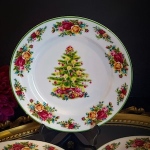 Puede incluir: Plato navideño de cerámica blanca con borde verde y una imagen central de un árbol de Navidad decorado. El plato está adornado con un motivo floral de rosas rojas y amarillas. Una estrella corona el árbol y hay regalos en la base.