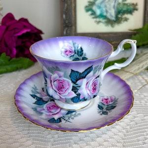May include: A purple teacup and saucer set with a floral design featuring white roses and green leaves. The set has a gold trim around the edges.