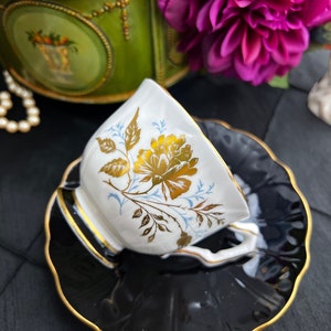 May include: A white teacup with a gold floral design sits on a black saucer with a gold rim. The saucer has a scalloped edge.