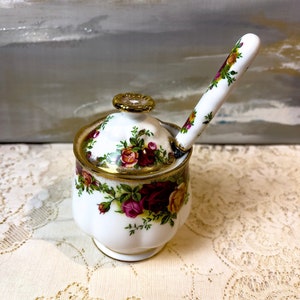 May include: A white ceramic sugar bowl with a lid and spoon, adorned with a floral rose pattern in shades of red, yellow, and green. The rim and handle are trimmed with gold. The spoon rests in the bowl.