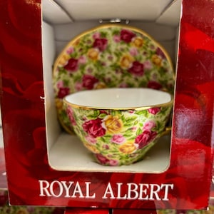 May include: A red box with a white teacup and saucer set inside. The teacup and saucer are decorated with a floral pattern of red, yellow, and pink roses. The box has the words "Royal Albert" printed on the front.
