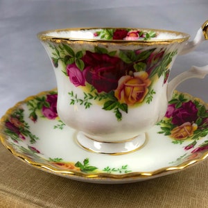May include: A white teacup and saucer set with a floral rose pattern in shades of red, pink, and yellow, accented with green leaves. The rims are trimmed with gold. The teacup sits on a matching saucer.