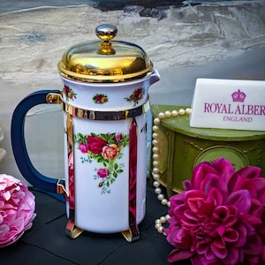 Może przedstawiać: Biały porcelanowy zaparzacz do kawy typu french press ze złotą pokrywką i niebieskim uchwytem. Zaparzacz zdobi kwiatowy wzór z czerwonymi różami i zielonymi liśćmi. Zaparzacz stoi na czarnym tle z naszyjnikiem z pereł i różowymi kwiatami.