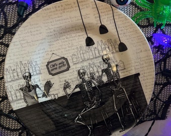 May include: White ceramic plate with a black and white illustration of skeletons at a bar. The plate has a black and white script pattern and the text "We only serve spirits".