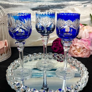 Puede incluir: Tres copas de vino de cristal azul cobalto con intrincados patrones tallados, exhibidas en una bandeja espejada con un borde tachonado de cristal. Las copas tienen tallos largos y están dispuestas en fila, reflejando la luz.