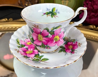 Puede incluir: Un juego de taza y platillo de té blanco con un patrón floral de rosas rosas y ribete dorado. El platillo tiene un borde festoneado. La taza de té tiene un asa y las palabras "Royal Albert England" están impresas en una tarjeta separada.