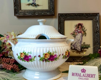 May include: White porcelain soup tureen with a lid, adorned with a floral pattern of yellow and purple roses. The tureen has gold trim and a pedestal base. A Royal Albert England sign is in front.