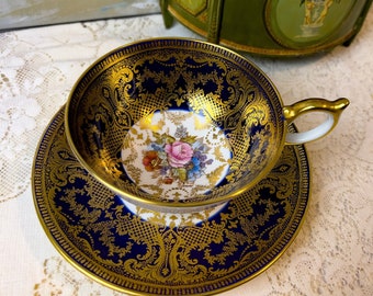 Può includere: Un set da tè composto da tazza e piattino con un motivo floreale. La tazza e il piattino hanno un bordo dorato e intricati motivi dorati su uno sfondo blu scuro.