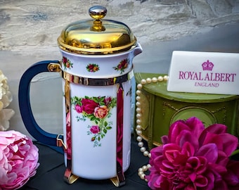 May include: A white porcelain French press coffee maker with a gold lid and blue handle. The coffee maker features a floral pattern with red roses and green leaves. The coffee maker is sitting on a black surface with a string of pearls and pink flowers.
