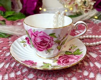 Può includere: Una tazza da tè e un piattino bianchi con un motivo floreale di rose rosa. La tazza da tè ha un bordo dorato e si trova su un sottopentola in pizzo rosa. Una collana di perle è all'interno della tazza da tè.