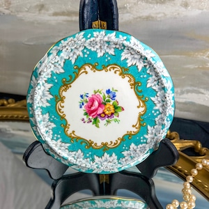 May include: A vintage trinket tray, round in shape, with a teal background and floral design. The tray has a white central area with a bouquet of pink, purple, and yellow flowers. The rim is decorated with white floral patterns and gold accents. It is on a black stand.