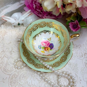 May include: A vintage teacup and saucer set in a soft mint green with gold filigree detailing. The teacup interior features a floral design with pink, purple, and yellow flowers. A pearl necklace and decorative white ceramic bunnies are also in the image.