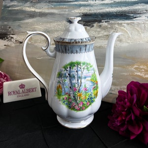 May include: White porcelain teapot with a floral design featuring a lake and birch trees. The teapot has a gold trim and a lid with a floral design. The teapot is made by Royal Albert England.