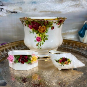 May include: A set of three miniature teacups and saucers featuring a floral pattern. The cups are white with a gold trim and feature a floral design with red, yellow, and pink roses. The saucers are white with a gold trim and feature a floral design with red, yellow, and pink roses.