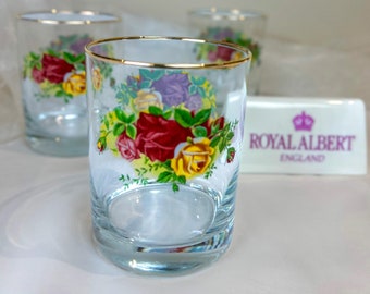 May include: Three clear glass tumblers with a gold rim and floral pattern. The tumblers feature a floral design with red, yellow, and purple roses. The tumblers are sitting on a white surface. The text "ROYAL ALBERT ENGLAND" is visible on a white rectangular surface in the background.